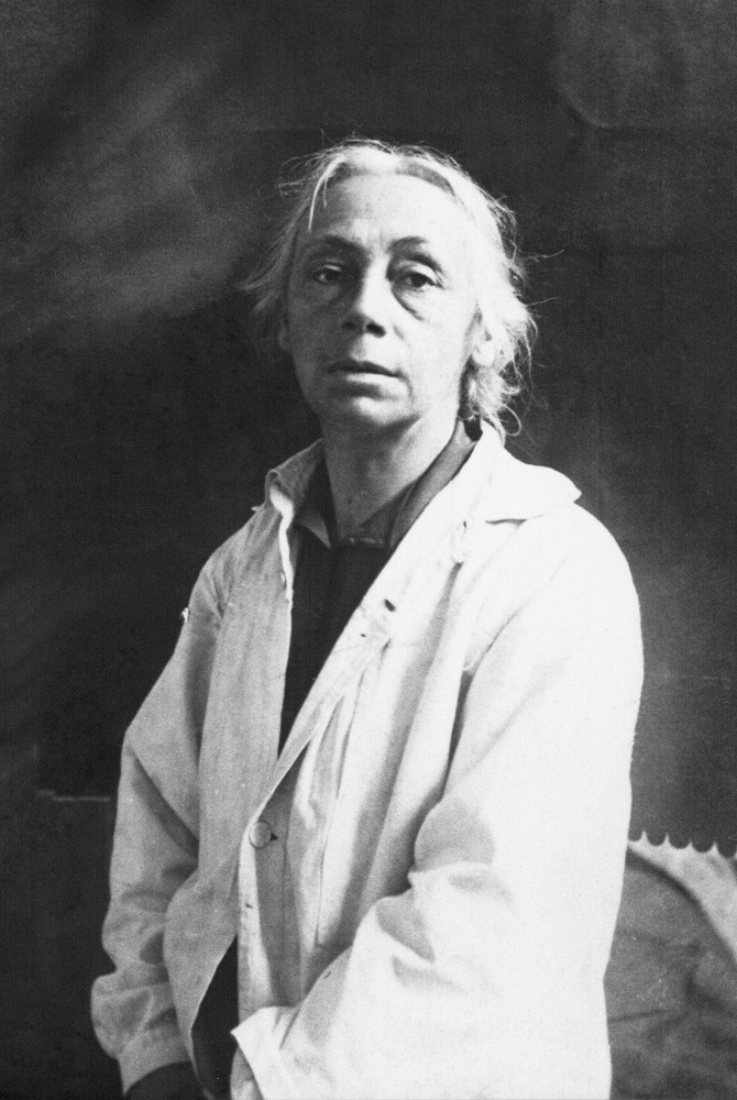 This black-and-white portrait depicts Käthe Kollwitz around 1920, standing in a frontal half-length shot against a dark, indistinct background. The artist, aged around 53, is wearing a light-coloured, simple smock, which is slightly open at the chest, revealing a darker undershirt. Her grey hair is parted at the top of her head and loosely swept back at the sides, with individual strands framing her face. Her gaze is directed straight at the camera and appears serious. Photographer unknown. A reproduction print of this photograph from the artist’s estate is held in the archives of the Kollwitz Museum Köln, Inv.: No. F 01.b-12.