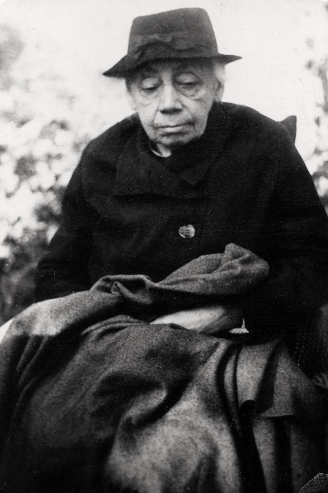 This black-and-white portrait, dating from around 1944–45, shows the elderly Käthe Kollwitz seated in a pensive pose, her upper body filling the centre of the frame. She wears a dark, simple hat and a heavy coat, whilst her furrowed face, with eyes almost closed, radiates a deep, meditative calm. Her hands rest unseen on her lap beneath a heavy dark blanket. It is perhaps the last photograph of the artist, taken in Moritzburg. The photographer is Hartmut Reiche. A reproduction print from the artist’s estate is held in the archives of the Kollwitz Museum in Köln, Inv. No. F-01.a-49. 