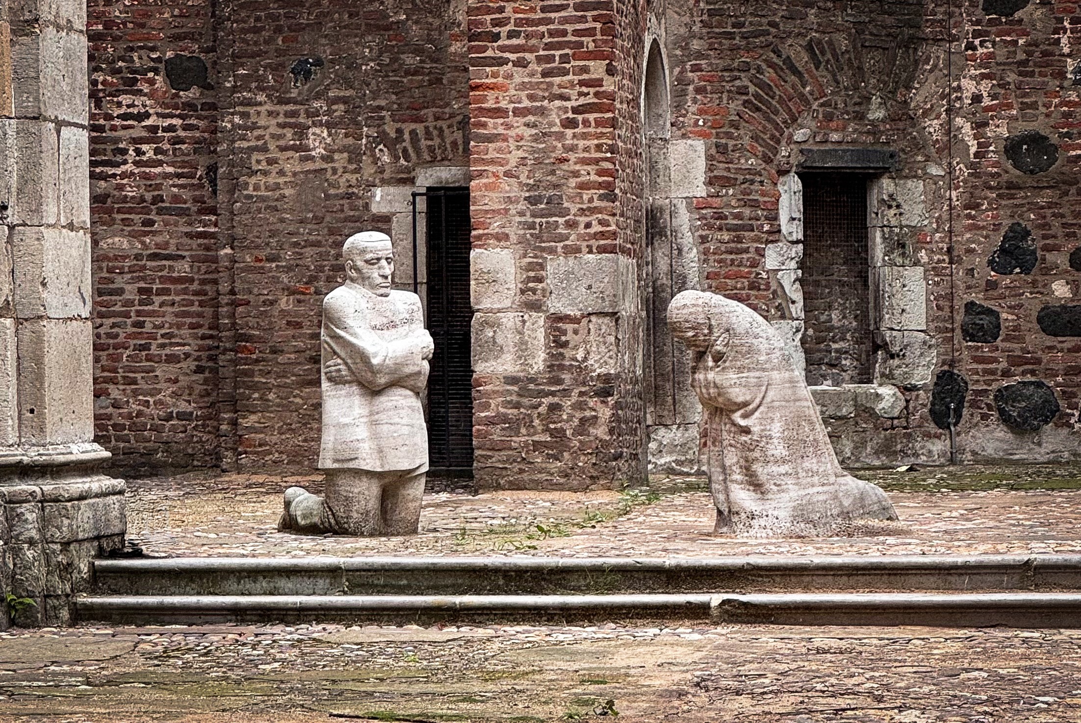 This photograph by Christian Nitz shows Käthe Kollwitz’s Memorial »Grieving Parents«, which is situated in the open interior of the ruins of the Old St Alban’s Church in Cologne. Amidst the weathered brick walls, two life-size, block-like stone figures kneel facing each other in silent grief: on the left, the father stands motionless, his gaze fixed straight ahead and his arms crossed tightly across his chest, whilst on the right, the mother, deeply bowed and lost in her own thoughts, mourns their son.
