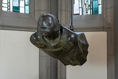 This photograph by Chris Franken shows the bronze sculpture »Der Schwebende« (The Grieving Angel) by Ernst Barlach — the second cast after the original working model of 1939 — in its present location in the Antoniterkirche in Cologne, seen frontally and turned to the left. The life-size bronze figure, hovering horizontally, hangs freely in the air from two chains, its straight body and arms crossed over the chest radiating a deep serenity. The face of the sculpture has closed eyes and seems absorbed in itself, while the flowing form of the robe reinforces the impression of a gentle flight.