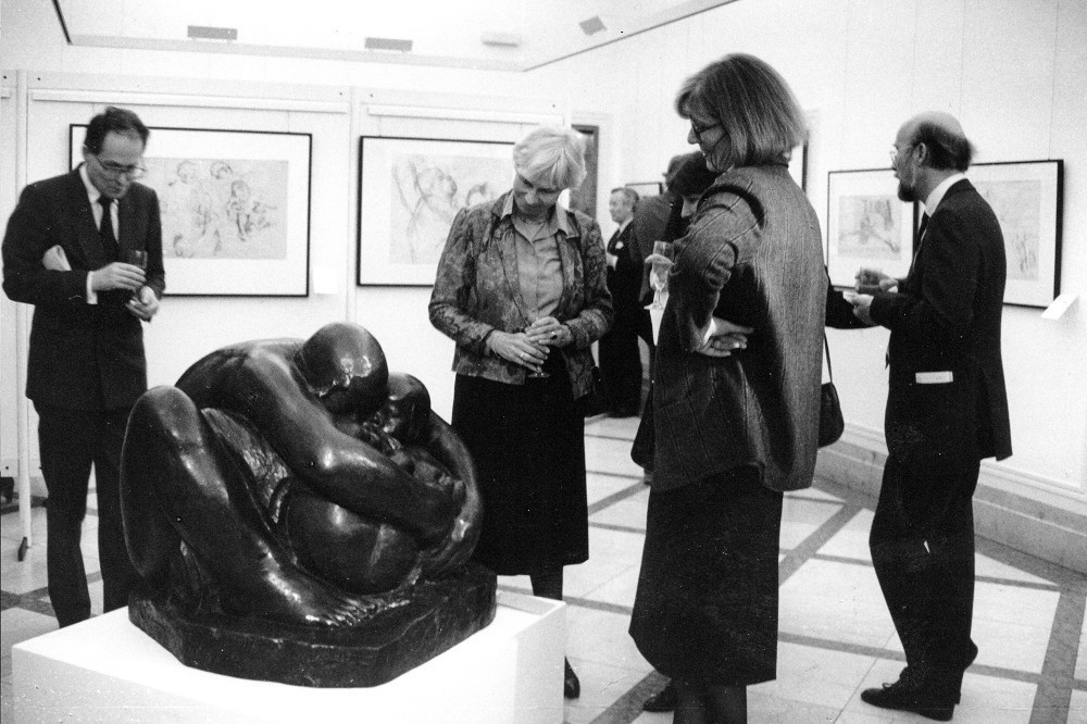 The black-and-white photograph shows a view of the museum’s first exhibition rooms, located on the former executive floor of the Kreissparkasse Köln, in 1986: several women and men are viewing works by Kollwitz, with the large bronze sculpture »Mother with Two Children« in the centre. 