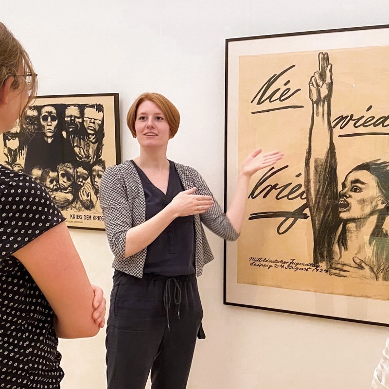 In the brightly lit exhibition rooms of the Kollwitz Museum in Cologne, an art educator uses sweeping hand gestures to explain Käthe Kollwitz’s famous anti-war poster “Never Again War” to two visitors who are listening intently. The artwork takes center stage, featuring an expressive black-and-white charcoal drawing of a person passionately demanding “Never Again War” with her mouth wide open and her hand raised in a vow.