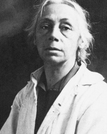 Das Schwarz-Weiß-Porträt zeigt Käthe Kollwitz um 1920, stehend, in einer frontalen Halbkörperaufnahme vor einem dunklen, unbestimmten Hintergrund. Die etwa 53-jährige Künstlerin trägt einen hellen, schlichten Arbeitskittel, der über der Brust leicht geöffnet ist und den Blick auf ein dunkleres Untergewand freigibt. Ihr graues Haar ist am Oberkopf gescheitelt und an den Seiten locker zurückgenommen, wobei einzelne Strähnen ihr Gesicht umrahmen. Ihr Blick ist direkt in die Kamera gerichtet und wirkt ernst. Fotograf:in unbekannt. Ein Reproabzug dieses Fotos aus dem Nachlass der Künstlerin befindet sich im Archiv des Kollwitz Museum Köln, Inv.: Nr. F 01.b-12.