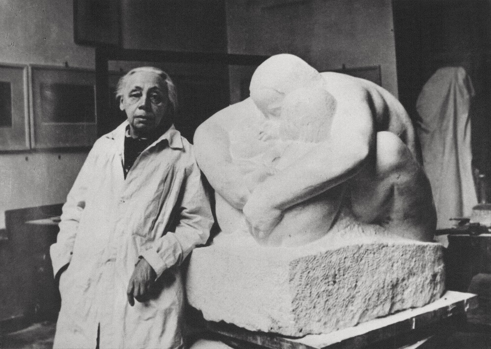 Dieses Schwarz-Weiß-Foto, entstanden wohl 1938, zeigt Käthe Kollwitz in ihrem Atelier in der Berliner Klosterstraße. Die etwa 71-jährige steht links neben der Gipsform ihrer Plastik »Mutter mit zwei Kindern«. Sie trägt einen langen, hellen Arbeitskittel und blickt mit ernstem Gesichtsausdruck direkt in die Kamera, während ihre linke Hand locker auf dem Sockel Ihres Werks ruht. Die überlebensgroße Skulptur zeigt eine Mutterfigur, die schützend über zwei kleine Kinder gebeugt ist und diese fest in ihren Armen umschließt. Im Hintergrund sind die schlichten Wände des Ateliers mit einigen gerahmten Bildern und anderen verhüllten Objekten zu erkennen, was die Arbeitsatmosphäre unterstreicht. Fotograf:in unbekannt. Ein Reproabzug dieses Fotos aus dem Nachlass der Künstlerin befindet sich im Archiv des Kollwitz Museum Köln, Inv.: Nr. F 01.a-37.