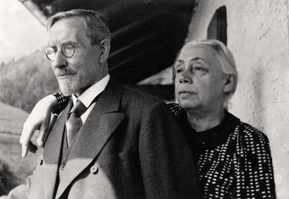 Das Schwarz-Weiß-Foto aus dem Jahr 1935 zeigt das Ehepaar Käthe und Karl Kollwitz als Brustporträt, aufgenommen während ihres Urlaubs in Bad Reichenhall, vor ihrem Ferienhaus im Freien. Der elegant gekleidete Karl Kollwitz steht im Vordergrund, trägt Brille, Krawatte und einen dunklen Mantel, während er nachdenklich in die Ferne blickt. Käthe Kollwitz steht leicht versetzt hinter ihm. Sie trägt ein dunkles Kostum mit weißem Muster und hat ihren Arm vertraut auf seine Schulter gelegt. Sie schaut mit einem ernsten, gefassten Gesichtsausdruck in die gleiche Richtung. Fotograf:in unbekannt. Ein Reproabzug dieses Fotos aus dem Nachlass der Künstlerin befindet sich im Archiv des Kollwitz Museum Köln, Inv.: Nr. F 01.b-31.