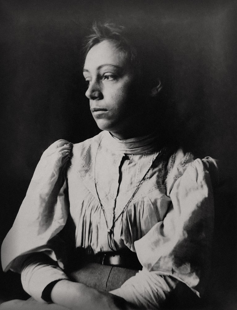 Eine Fotografie von Käthe Kollwitz, geborene Schmidt, im Alter von etwa 23 Jahren, sitzend vor dunklem Hintergrund. Sie blickt nach links und trägt eine weiße Bluse. Fotograf:in unbekannt. Das Vintage aus dem Nachlass von Mathilde Rüstow, eine Schenkung vonAlexandra Aichberger, befindet sich im Archiv des Kollwitz Museum Köln, Inv. Nr. F-01.a-03.