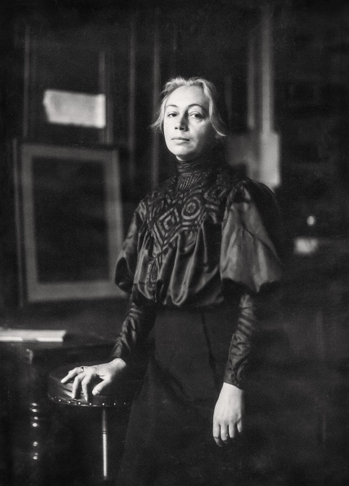 Eine Fotografie von Käthe Kollwitz aus dem Jahr 1906, stehend, im Hintergrund ihre Radierung »Carmagnole« von 1901. Die 39-jährige blickt ihre Betrachter:innen direkt an, stützt ihre rechte Hand auf ein hockerähnliches Möbel und trägt eine elegante dunkle, hochgeschlossene Bluse mit auffälligen Stickereien auf der Brust und voluminösen Ärmeln, die an der Schulter bauschig angesetzt sind und zum Handgelenk hin in eng anliegende Manschetten übergehen. Der Fotograf ist Philipp Kester. Ein Repro-Abzug aus dem Nachlass der Künstlerin befindet sich im Archiv des Kollwitz Museum Köln, Inv. Nr. F-01.a-06.