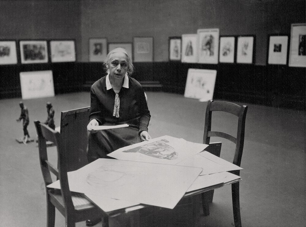 Das Schwarz-Weiß-Foto aus dem Jahr 1927 zeigt Käthe Kollwitz in einem Ausstellungssaal der Preußischen Akademie der Künste, Berlin. Die 60-jährige trägt ein schlichtes, dunkles Kostum mit weißem Kragen, unter dem ein helles Band ähnlich einer Kravatte gebunden ist.  
Sie sitzt vor einer improvisierten Arbeitsfläche, die aus zwei zusammengeschobenen Holzstühlen mit hohen Lehnen besteht. Die Sitzflächen bilden eine Art Tisch. Darauf liegen einige ihrer Werke, großformatige Papierbögen, von denen sie einen mit beiden Händen leicht anhebt, um ihn für die geplante Ausstellung auszuwählen. Im Hintergrund, an den Wänden des weiten Raums, sind bereits zahlreiche gerahmte Grafiken in einer Reihe aufgehängt. Ihr Blick ist direkt in die Kamera gerichtet und wirkt ernst und prüfend. Fotograf:in unbekannt. Ein Reproabzug dieses Fotos aus dem Nachlass der Künstlerin befindet sich im Archiv des Kollwitz Museum Köln, Inv.: Nr. F 01.e-01.