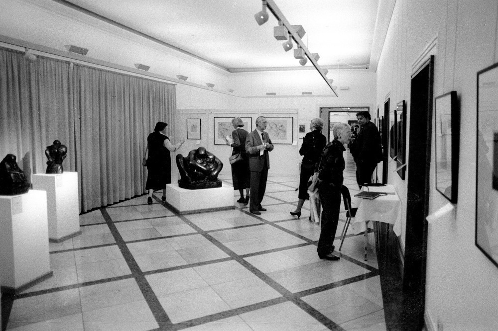 Das Schwarz-Weiß-Foto zeigt einen Blick in die ersten Ausstellungsräume des Museums auf der ehemaligen Vorstandsetage der Kreissparkasse Köln, 1986:  Mehrere Frauen und Männer betrachten Werke der Kollwitz, im Hintergund die große Bronzeplastik »Mutter mit zwei Kindern«. 