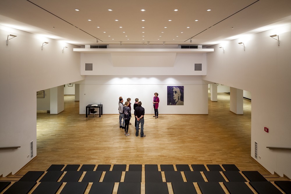 Das Farbfoto zeigt einen Blick in das Forum des Kollwitz Museum Köln im Jahr 2015: einen weiten, hellen Raum mit hellem Holzfußboden, an der Schauwand ein Kollwitz-Porträt, bearbeitet vom Künstler Herbert Döring-Spengler. Im Vordergrund eine Gruppe von Personen. 