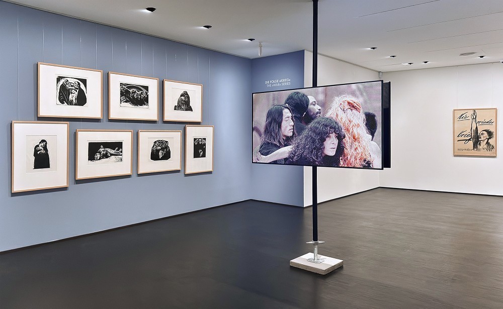 Das Foto zeigt einen Blick in die Sammlungspräsentation zur Wiedereröffnung des Kollwitz Museum Köln mit der ersten Ausstellungsintervention der Reihe »Bildstörung«: Zentral die Videoinstallation »Die Mütter« von Isaac Chong Wai, rechts die sieben Holzschnitte der Folge »Krieg«, rechts im Hintergrund das Plakat »Nie wieder Krieg« von Käthe Kollwitz. 