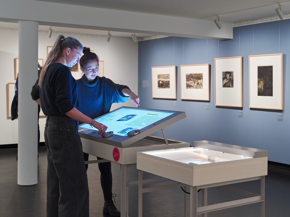 Das Foto zeigt einen Blick in die Sammlungsspräsentation zur Wiedereröffnung des Kollwitz Museum Köln 2025 mit der Medienstation zur Tiefdrucktechnik. Zwei Museumsbesucherinnen bedienen das pultartige, computergestütze Kunstvermittlungstool.