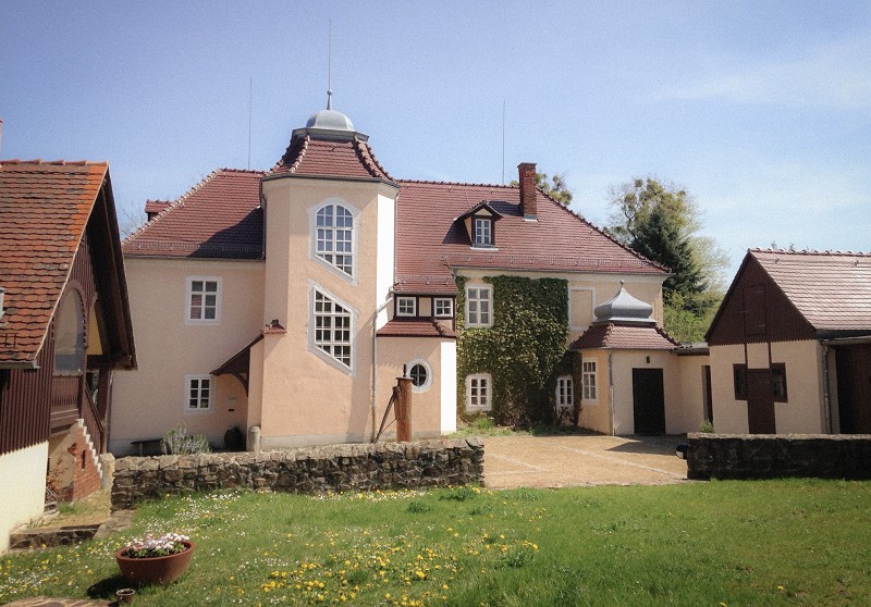 Das Bild zeigt den Rüdenhof in Moritzburg – den Sterbeort von Käthe Kollwitz. Ein helles, mehrstöckiges Gebäude mit einem markanten sechseckigen Treppenturm und rötlichen Ziegeldächern, das von einer niedrigen Natursteinmauer umgeben ist. Im Vordergrund erstreckt sich eine grüne Wiese mit gelben Löwenzahnblüten und einer kleinen Pflanzschale, während das Hauptgebäude teilweise mit Efeu bewachsen ist und von kleineren Nebengebäuden flankiert wird.